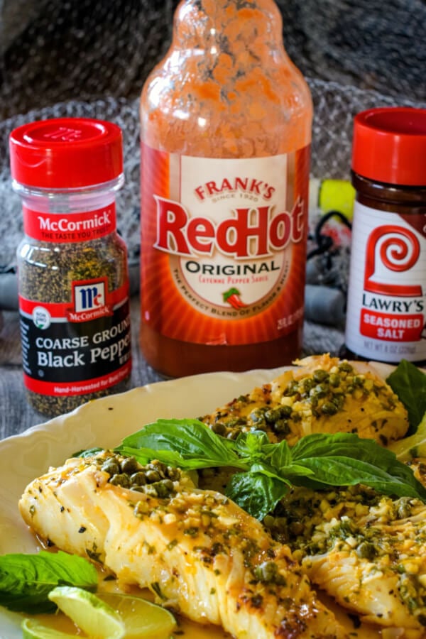 Grilled Halibut on a white plate with fresh basil sprig and lemons on the side ready to serve with bottle of red hot sauce, and jars of black pepper and seasoned salt. 