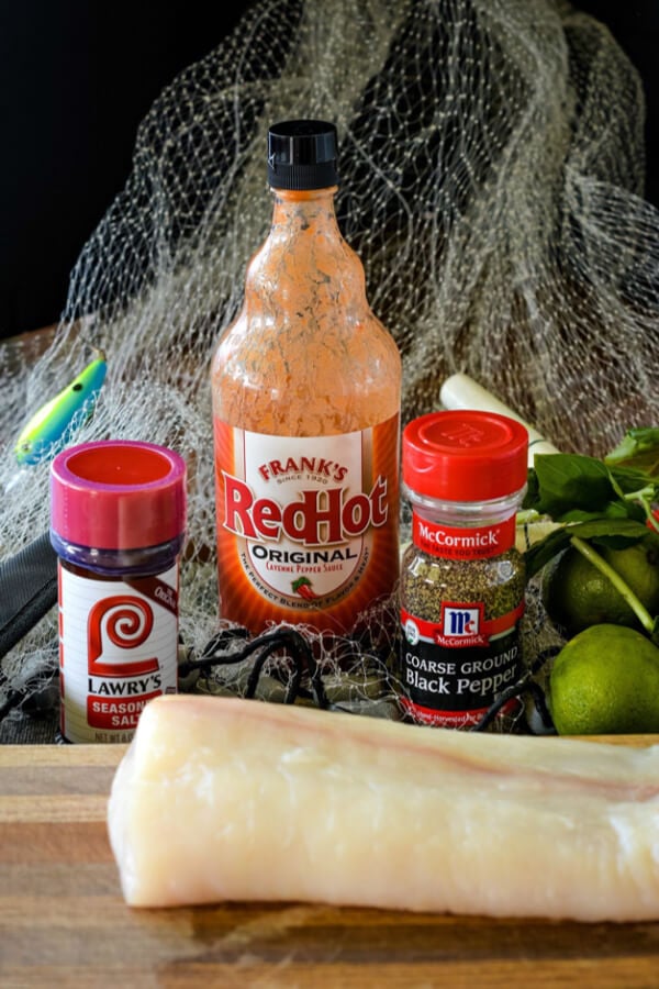 Raw halibut fillets on wood cutting board. A fishing net with a fishing lure and bobber in the net. Seasoned salt, hot sauce, and black pepper with a lime behind the fish. 