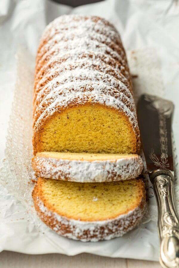 A long tube-like Polenta cake with white powdered sugar on top of a platter with silver serving knife on side. 