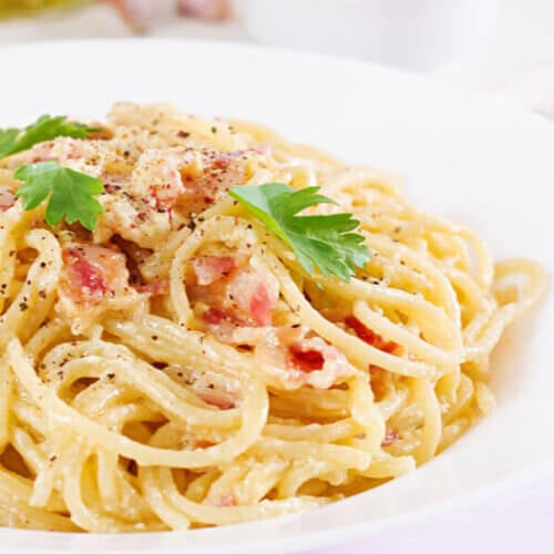 A white plate filled with egg sauce coated pasta with bits of pork on top and some parley leaves.