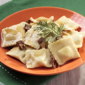 Mushroom ravioli in orange bowl with herb sprig on top and mushroom pieces surrounding the pasta. Green tablecloth underneath bowl.