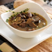 Beef mushroom soup in white bowl on white plate with sprig of fresh thyme.