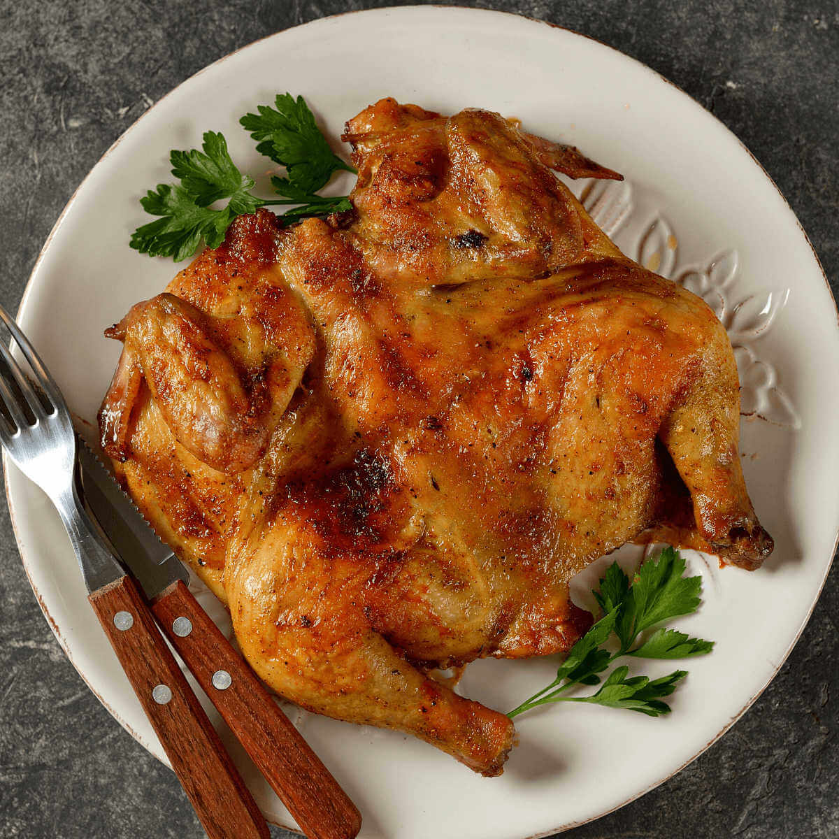 Grilled spatchcock chicken on white plate with knife and fork.