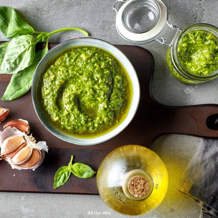 Green basil pesto in bowl with olive oil and garlic cloves on a wooden cutting board.
