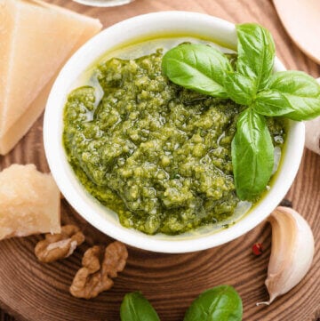 Bowl of green pesto surrounded by hard cheese, basil, walnuts and garlic.
