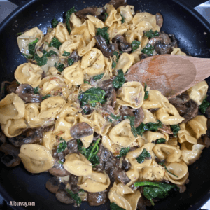 Skillet with wooden spoon stirring the saucy cheese tortellini with spinach, mushrooms and caramelized onions
