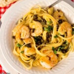 Lemon Caper shrimp on top of pasta in a white bowl on a red and white gingham tablecloth.