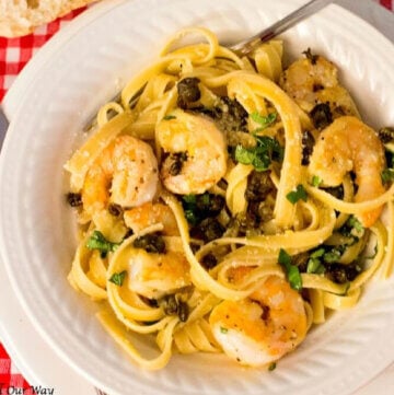 Lemon Caper shrimp on top of pasta in a white bowl on a red and white gingham tablecloth.