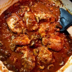 A large pan filled with braised chicken thighs covered in a red tomato wine sauce and mushrooms.