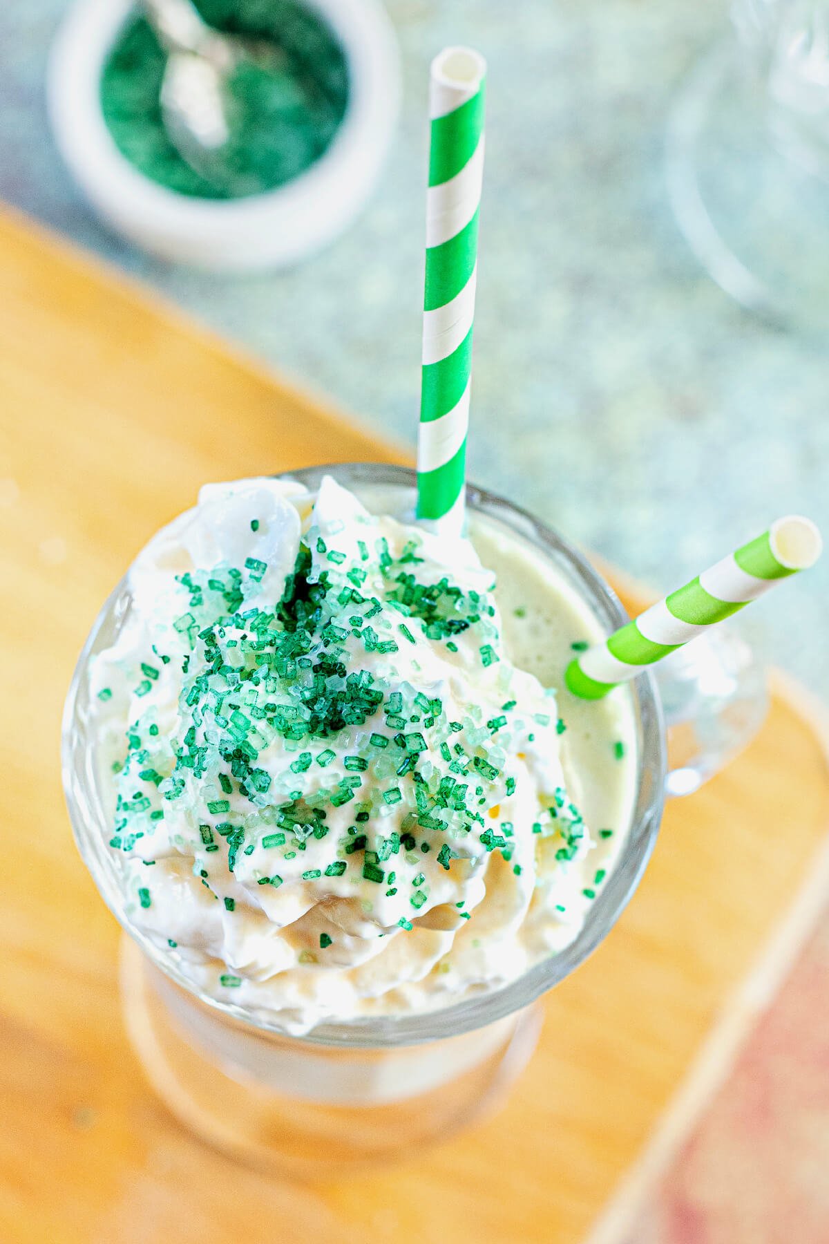 Overhead view of Bailey Iced Coffee with green sprinkles on top and two green striped straws stuck in the Irish Coffee glass mug. 