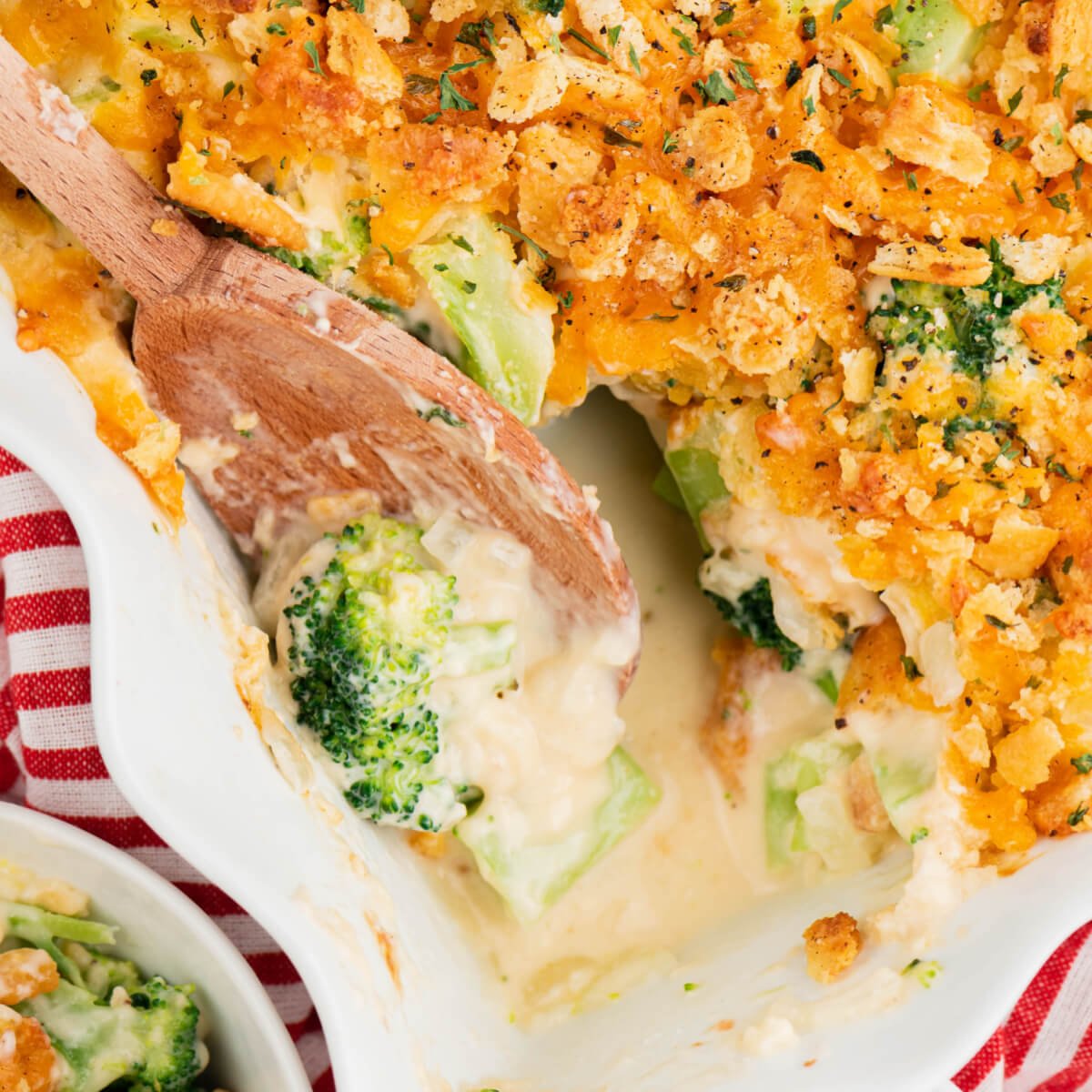 Cheesy Broccoli in a white casserole with a wooden spoon removing a portion.
