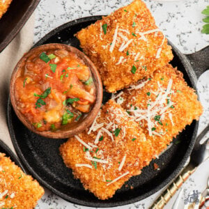 Three fried ravioli on a black serving dish with a bowl of spicy tomato dipping sauce next to them.