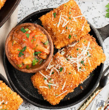 Three fried ravioli on a black serving dish with a bowl of spicy tomato dipping sauce next to them.