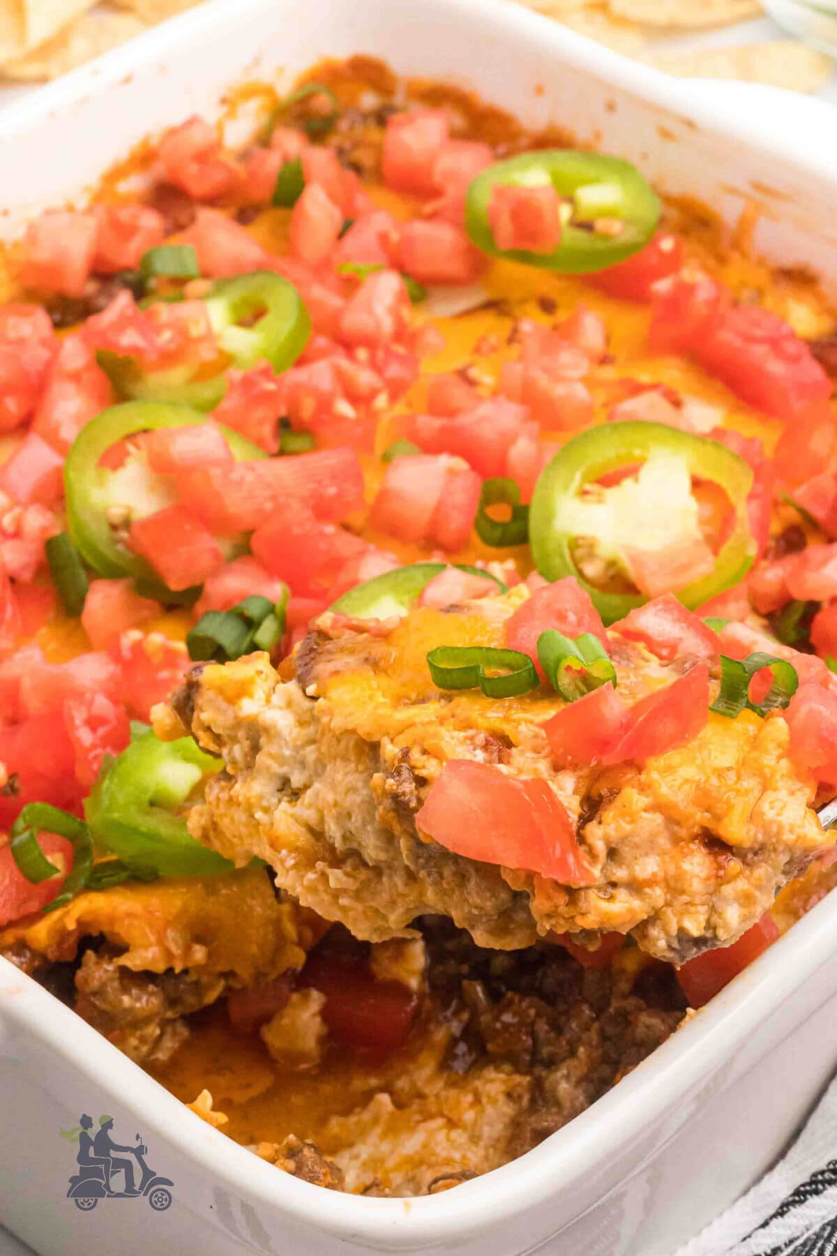 White casserole dish with Tex-Mex taco dip topped with tomatoes and jalapeños. 