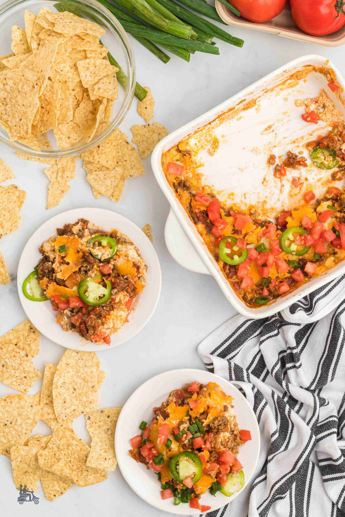 Image of two serving of baked taco dip on white plates with tortilla chips surrounding the plates. 