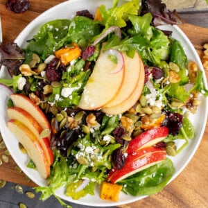 A white plate holding a tossed salad featuring autumn ingredients such as apples, sweet potatoes, pumpkin seeds, cranberries, and a mustard honey vinaigrette.