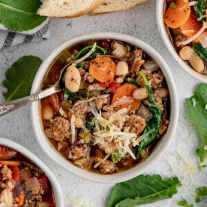 An overhead view of a bowl filled with Tuscan Sausage Kale soup on a white table and kale leaves sprinkled around the bowl.