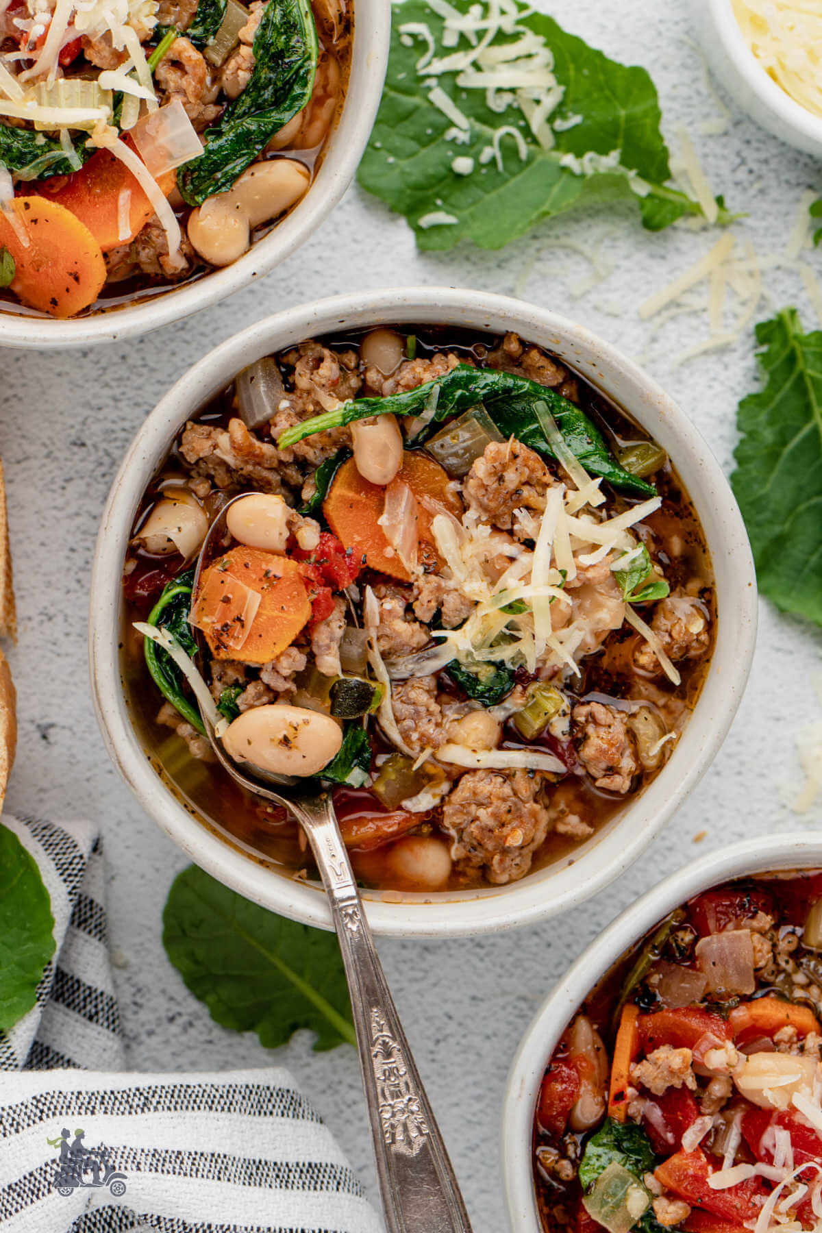 Three white bowls holding servings of Tuscan White Bean Soup made with sausage, tomatoes, carrots, and kale. 