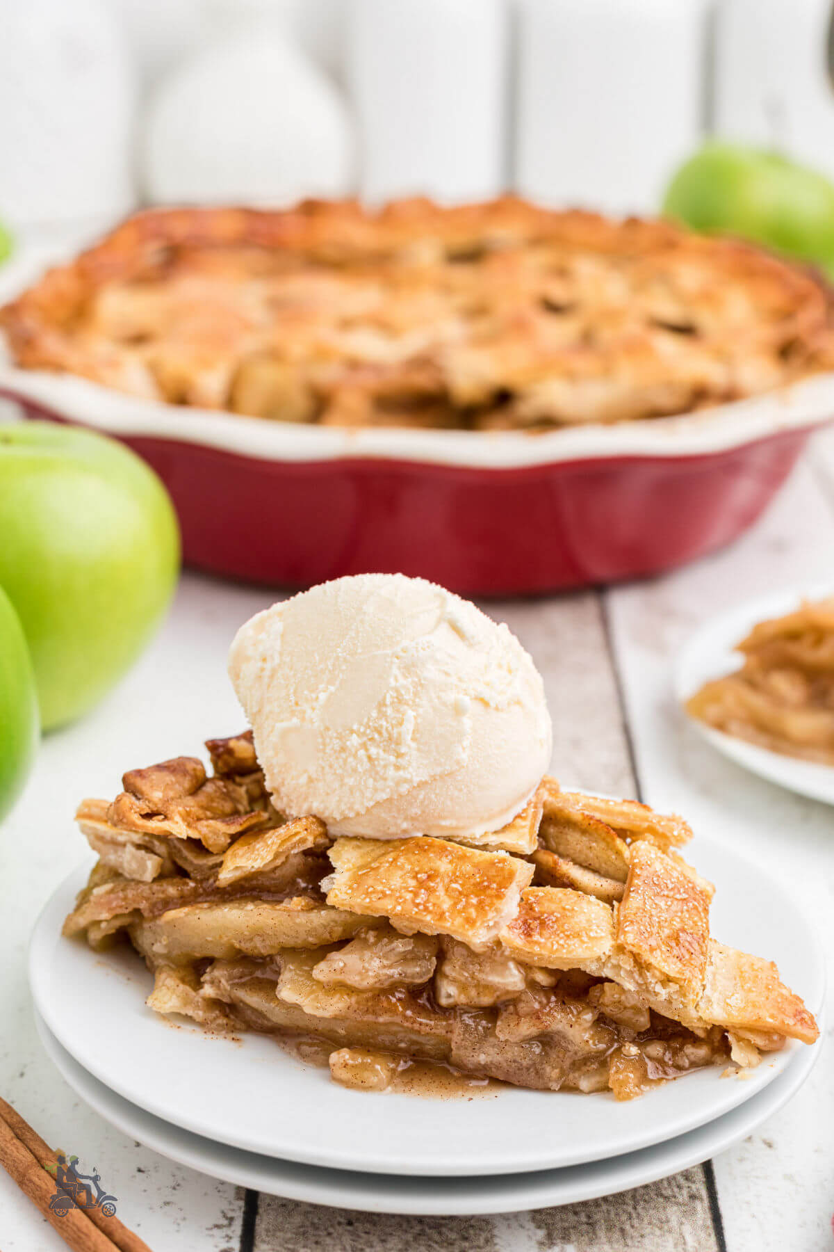 A slice of homemade American apple pie with a scoop of ice cream on top on a white plate with the rest of the dessert in a red pie plate in the background. 