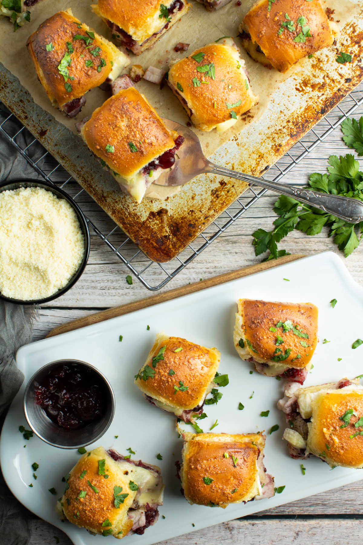 Cheesy Turkey sliders on baking sheet with some on a white serving plate with cranberry sauce in small bowl. 