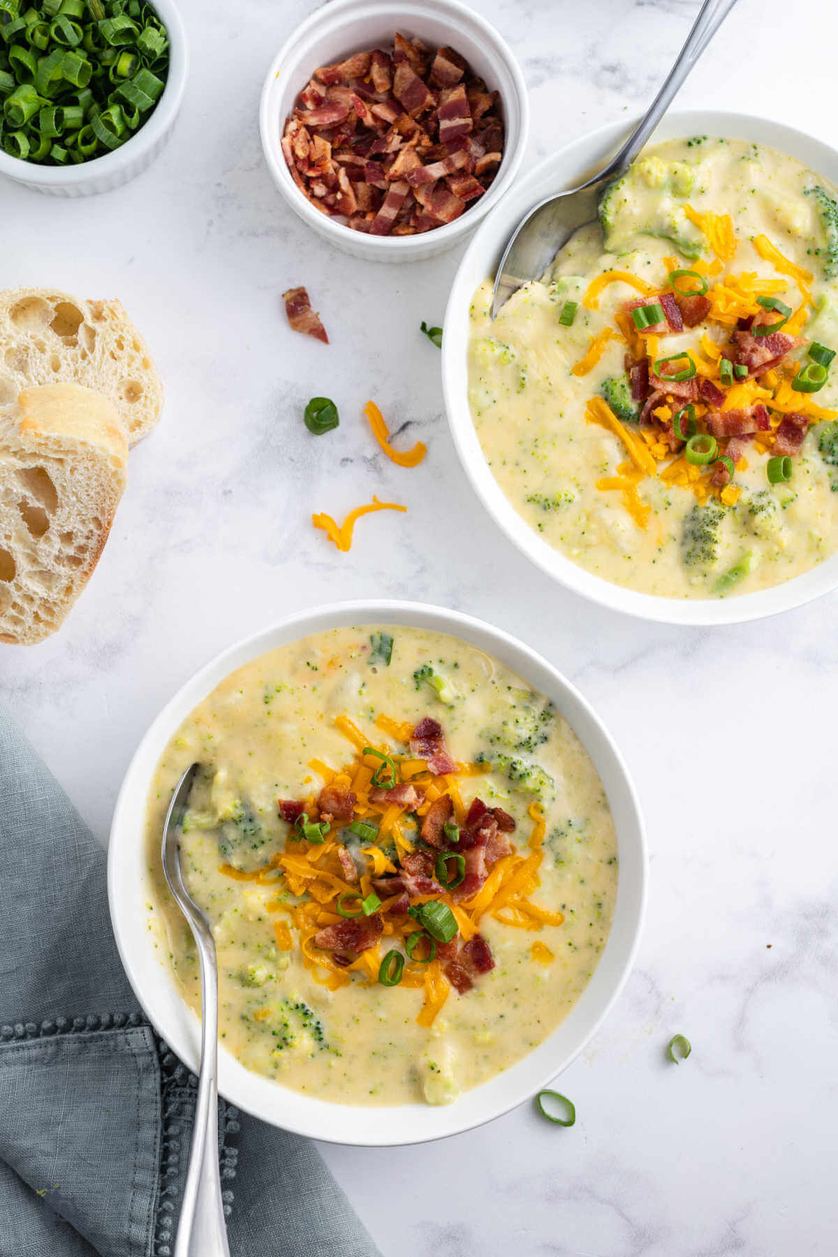 Two bowls of Potato Broccoli Soup with ciabatta bread on the counter and the soup garnishes of bacon and green onions. 