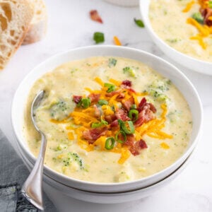 A white bowl filled with potato broccoli soup with cheddar cheese and bacon on top and a sprinkle of herbs on top.