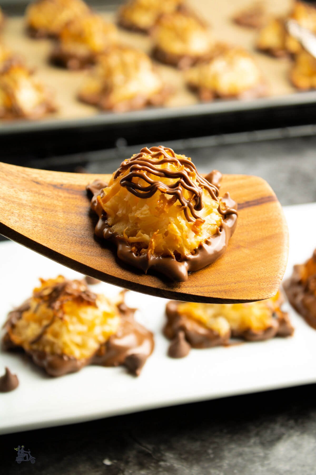 A wooden spatula holding a chocolate dipped coconut macaroon drizzled with melted chocolate on top. 
