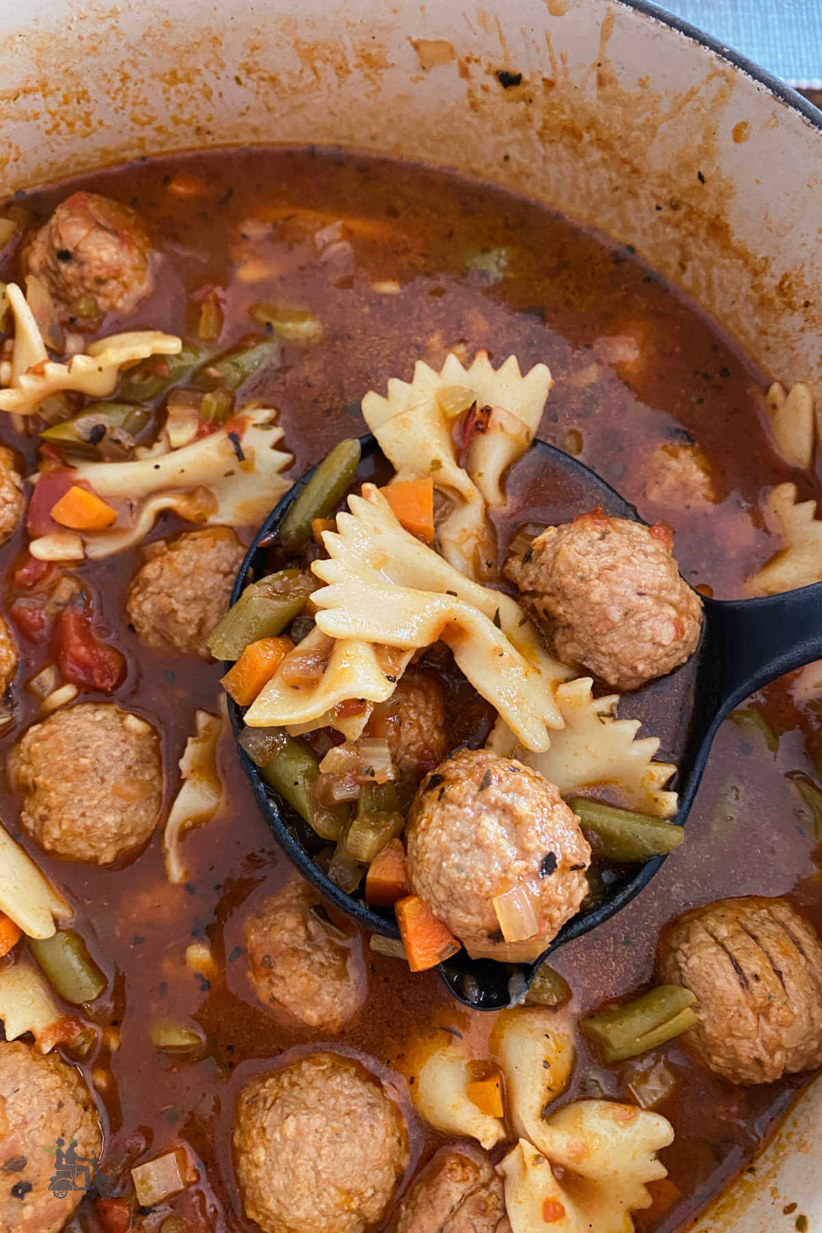 A Dutch Oven filled with beef meatball vegetable soup with bow tie pasta and a ladle holding up a scoopful. 