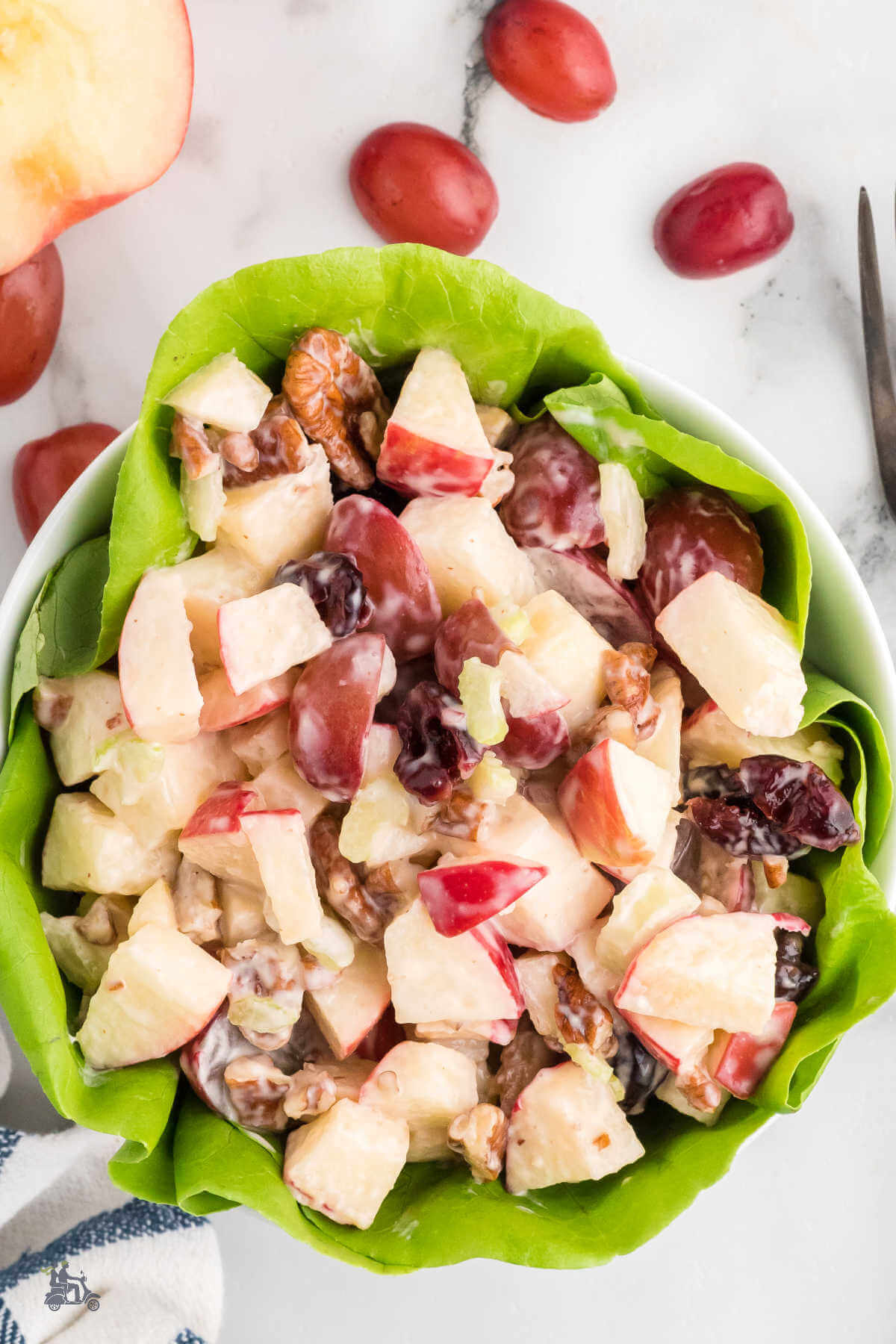 A lettuce lined white bowl filled with a Honeycrisp Harvest Apple Salad recipe with red grapes on a white counter and a half of a Honeycrisp apple. 