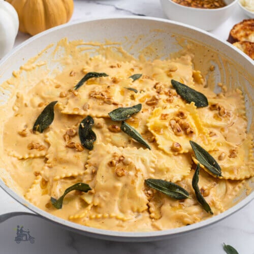 A large skillet filled with cheese ravioli with pumpkin cream sauce and topped with roasted chopped hazelnuts and fried sage leaves.