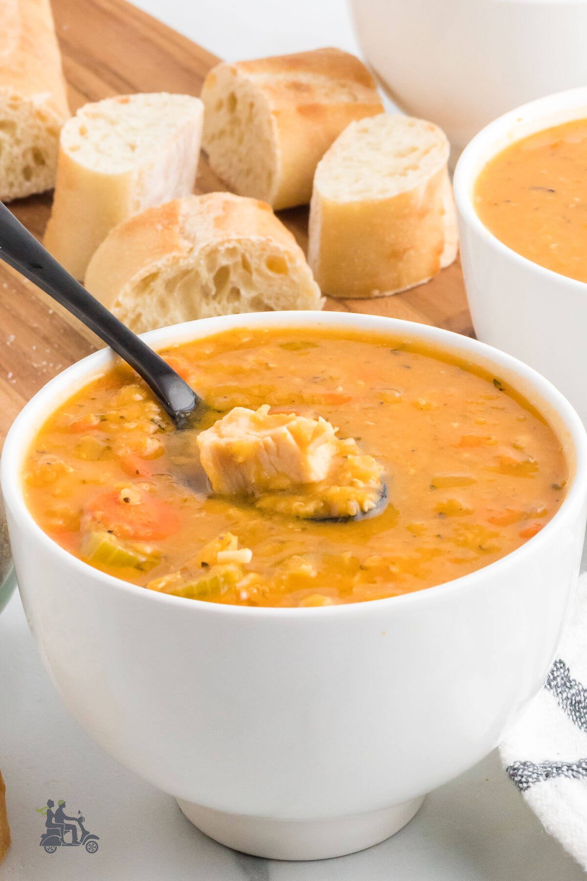 A white bowl filled with Chicken Lentil Soup and a soup dipping a spoonful with some sliced bread in the background. 