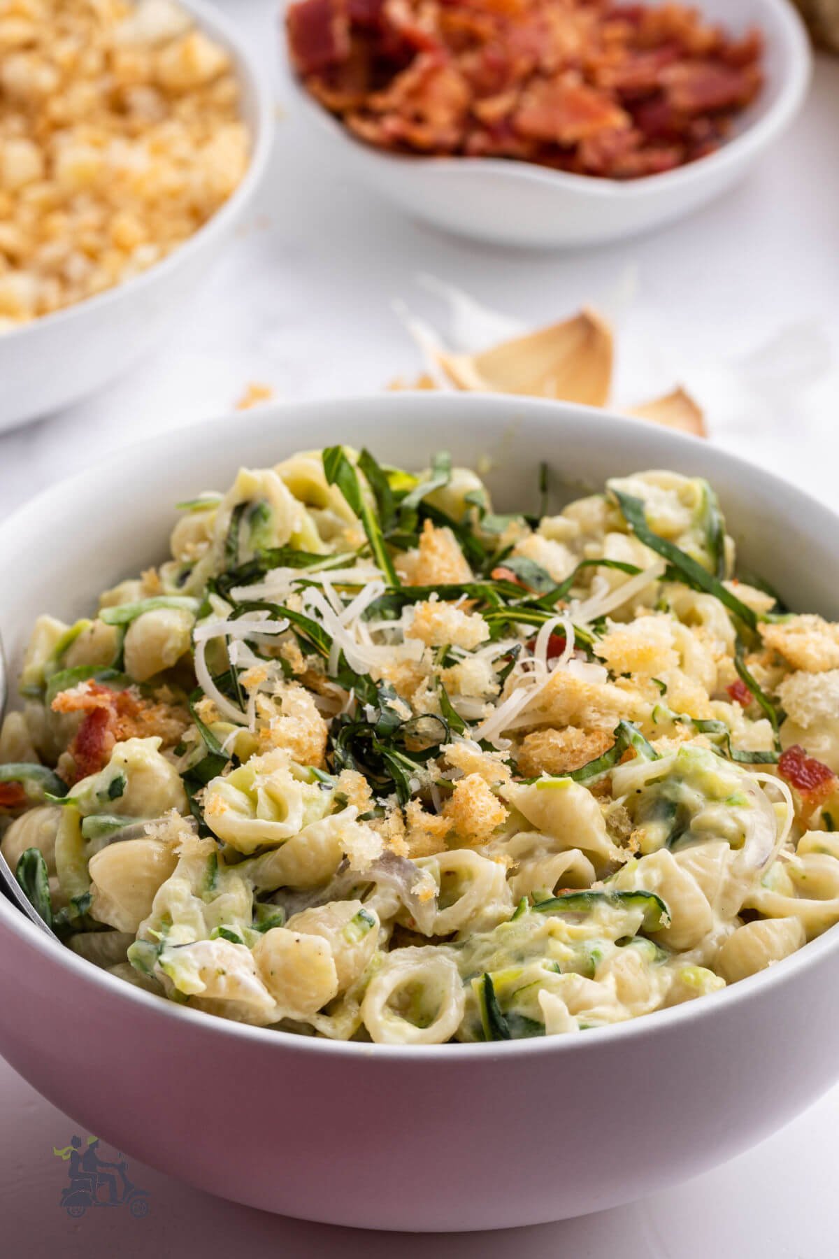 A white bowl filled with shell pasta coated with zucchini sauce and topped with bacon, parmesan, and buttered bread crumbs. 