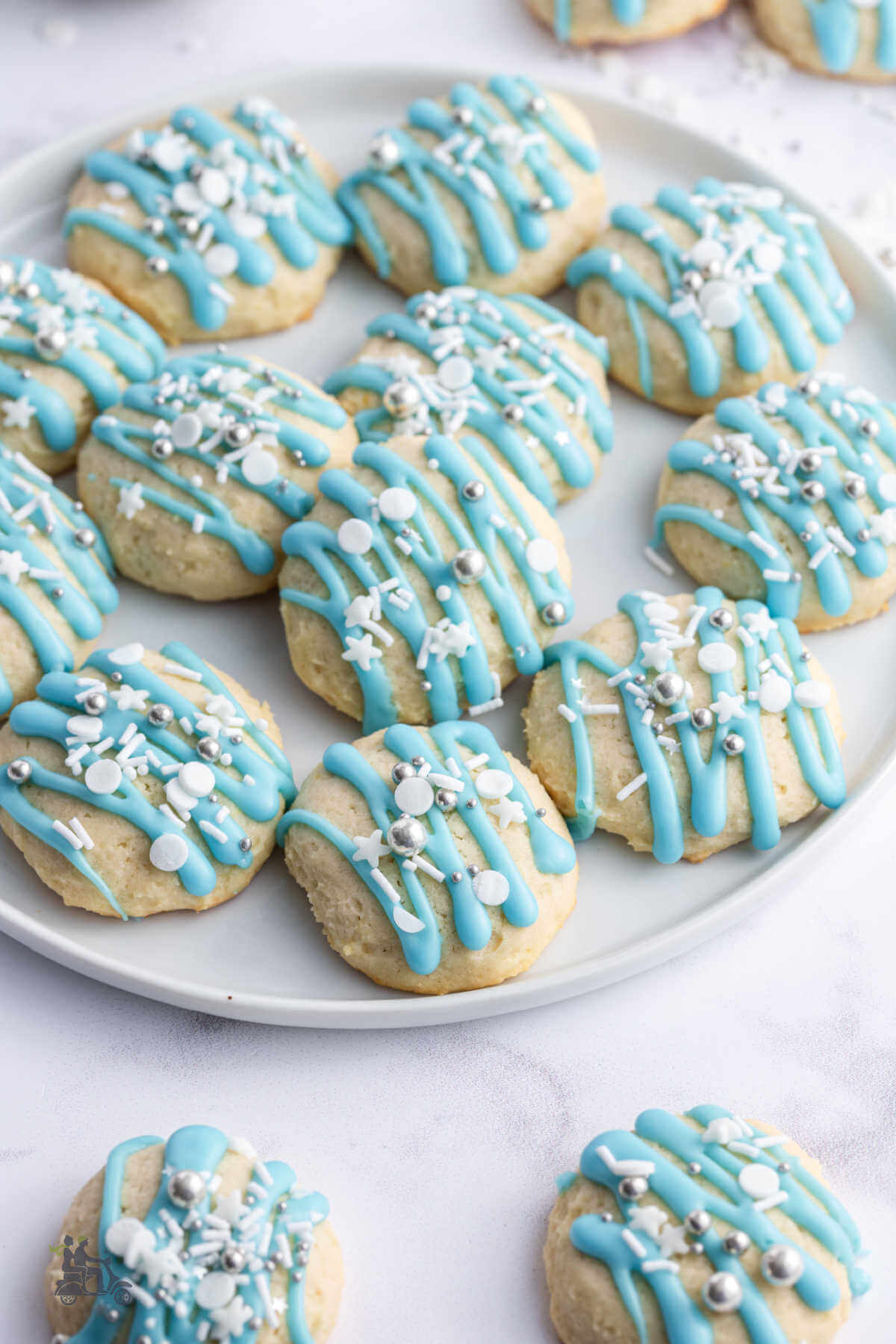 A white plate filled with Italia Lemon Ricotta Cookies drizzles with blue sugar icing and sprinkled with holiday sugars. 