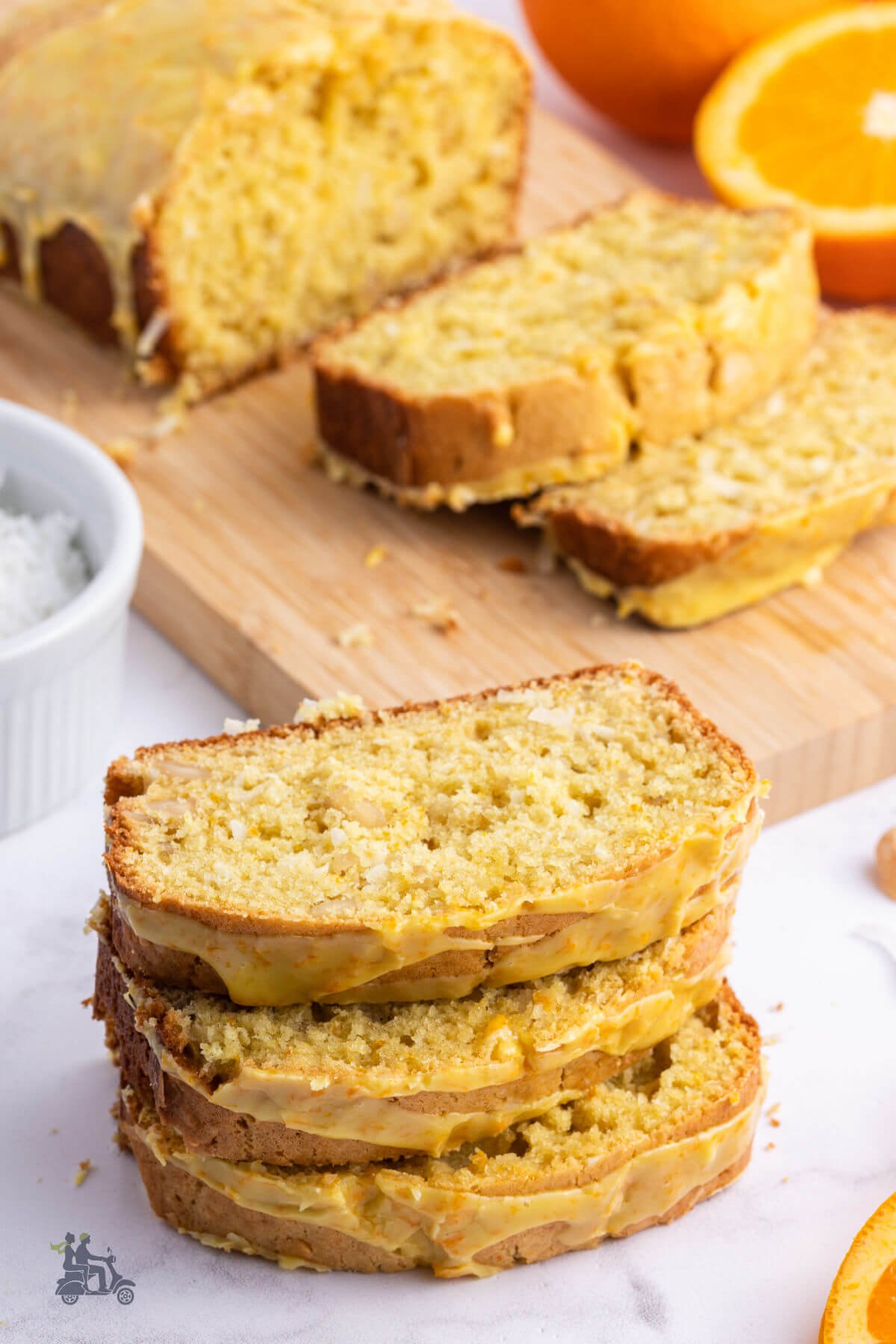 A loaf of Coconut Quick Bread with Orange Glaze on a wood cutting board with slices of the brad on the counter. 