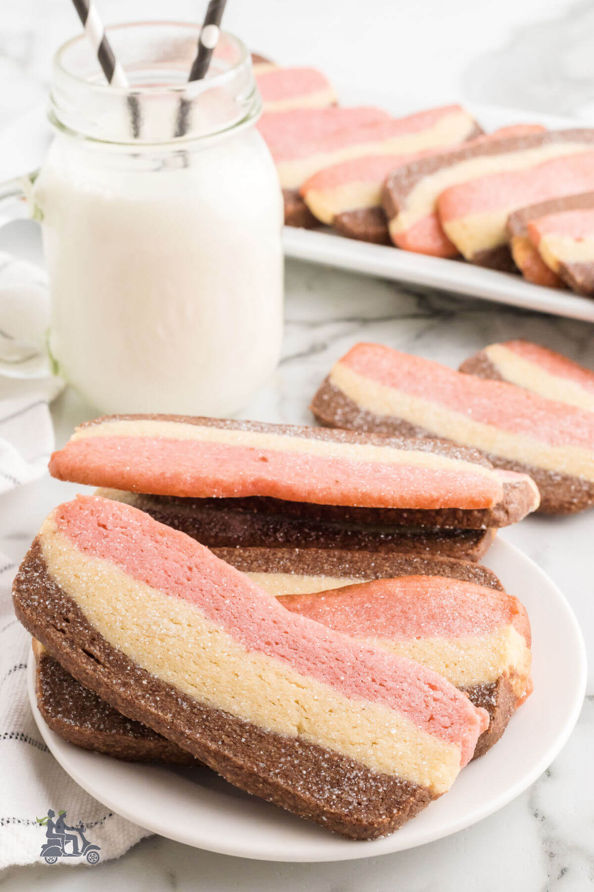 Tri-color pink, white, and chocolate Neapolitan Cookies on a white plate with a glass of milk on the counter. 