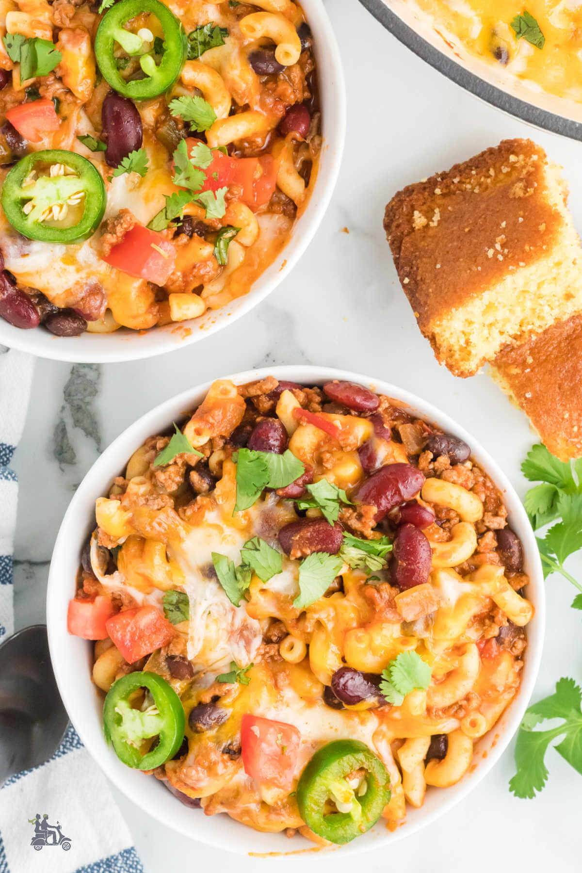 Overhead view of two white bowls filled with a Chili Mac and Cheese garnished with jalapenos and diced tomatoes with cornbread on the side. 