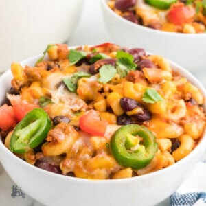 A close up of a bowl filled with ground beef chili mac and cheese with jalapeno slices and chopped tomatoes on top.