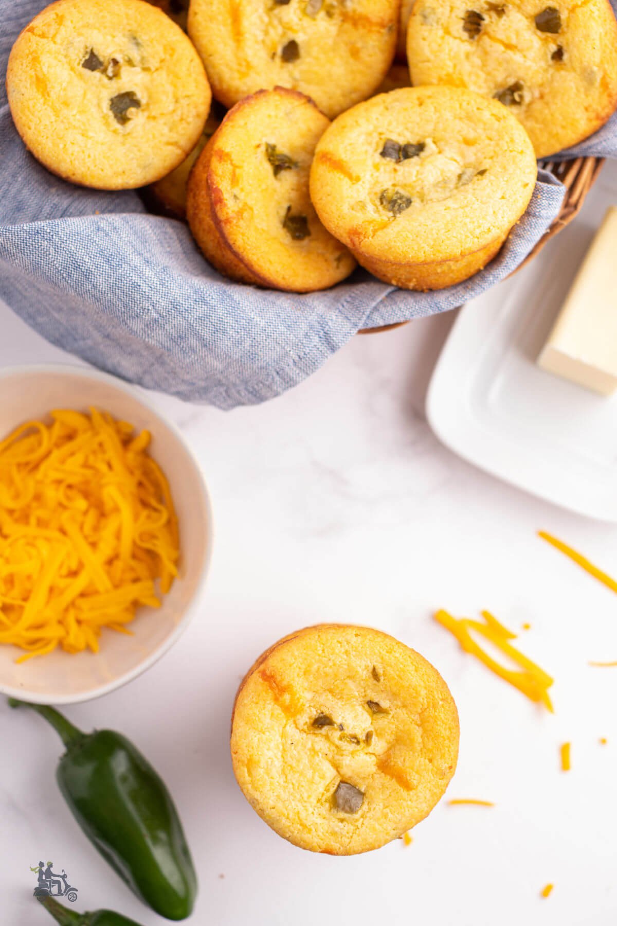 A basket of Jiffy corn bread muffins in a basket with a bowl of grated cheddar cheese and a dish of butter on the marble counter. 