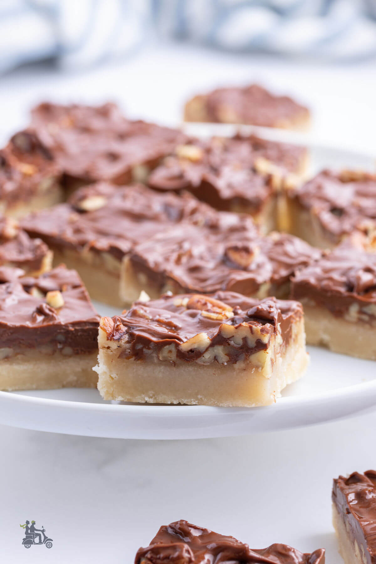 Turtle Pecan Bars with a milk chocolate topping on a white platter and surrounded by more caramel bars. 