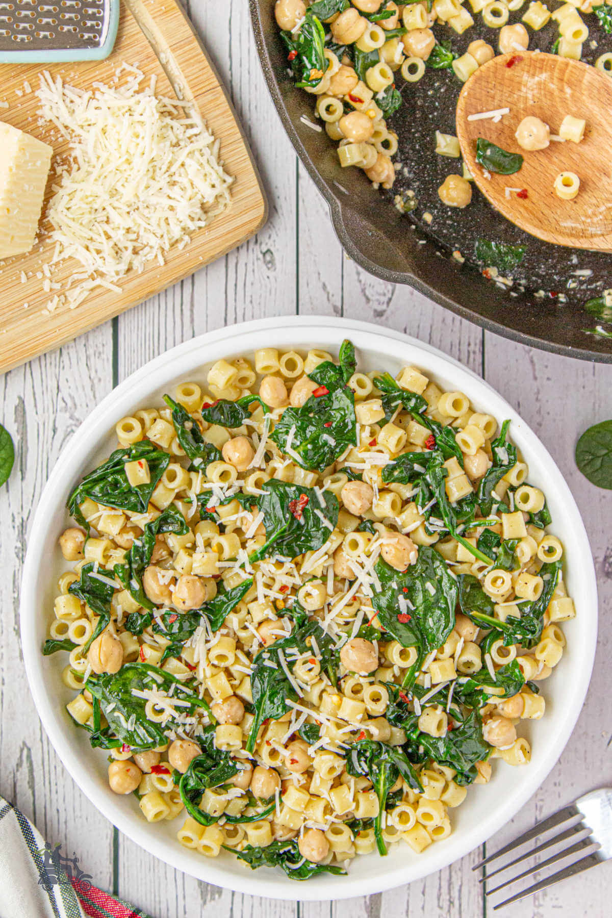 Chickpeas and pasta with spinach and red pepper flakes in a white ceramic bowl with a wood board holding pecorino romano grated cheese. 