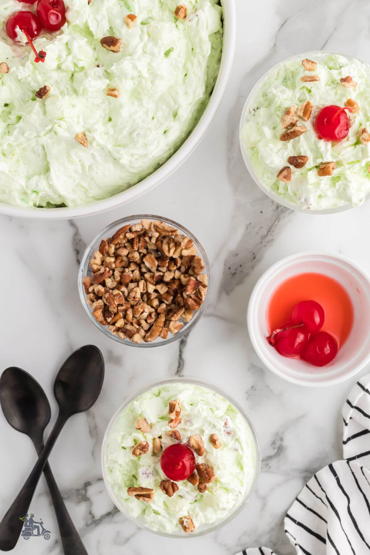 Overhead view of the classic watergate salad being served in glass goblets from a glass serving bowls. Maraschino cherries and chopped pecans on the side for garnish. 