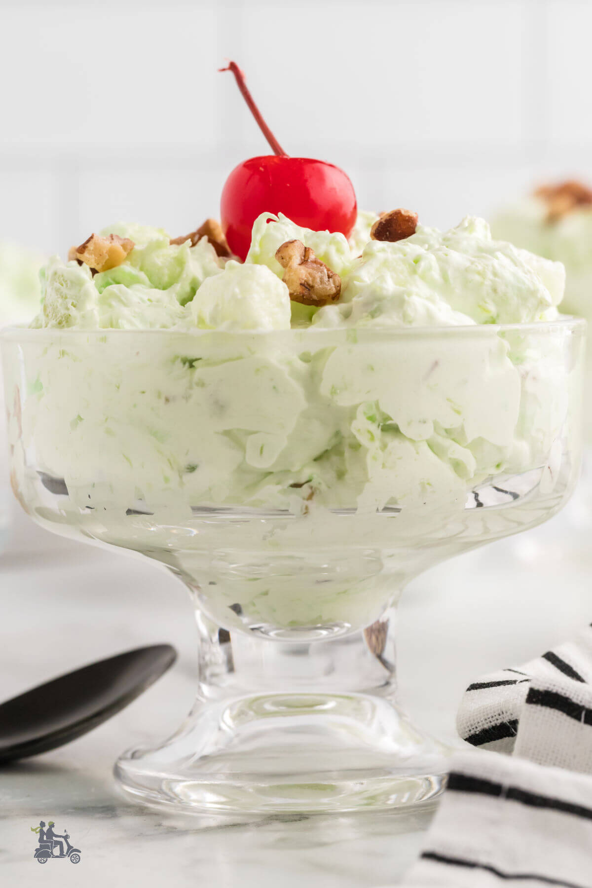 A side view of a glass goblet filled with pistachio fluff watergate salad with a maraschino cherry and chopped pecans on top. 