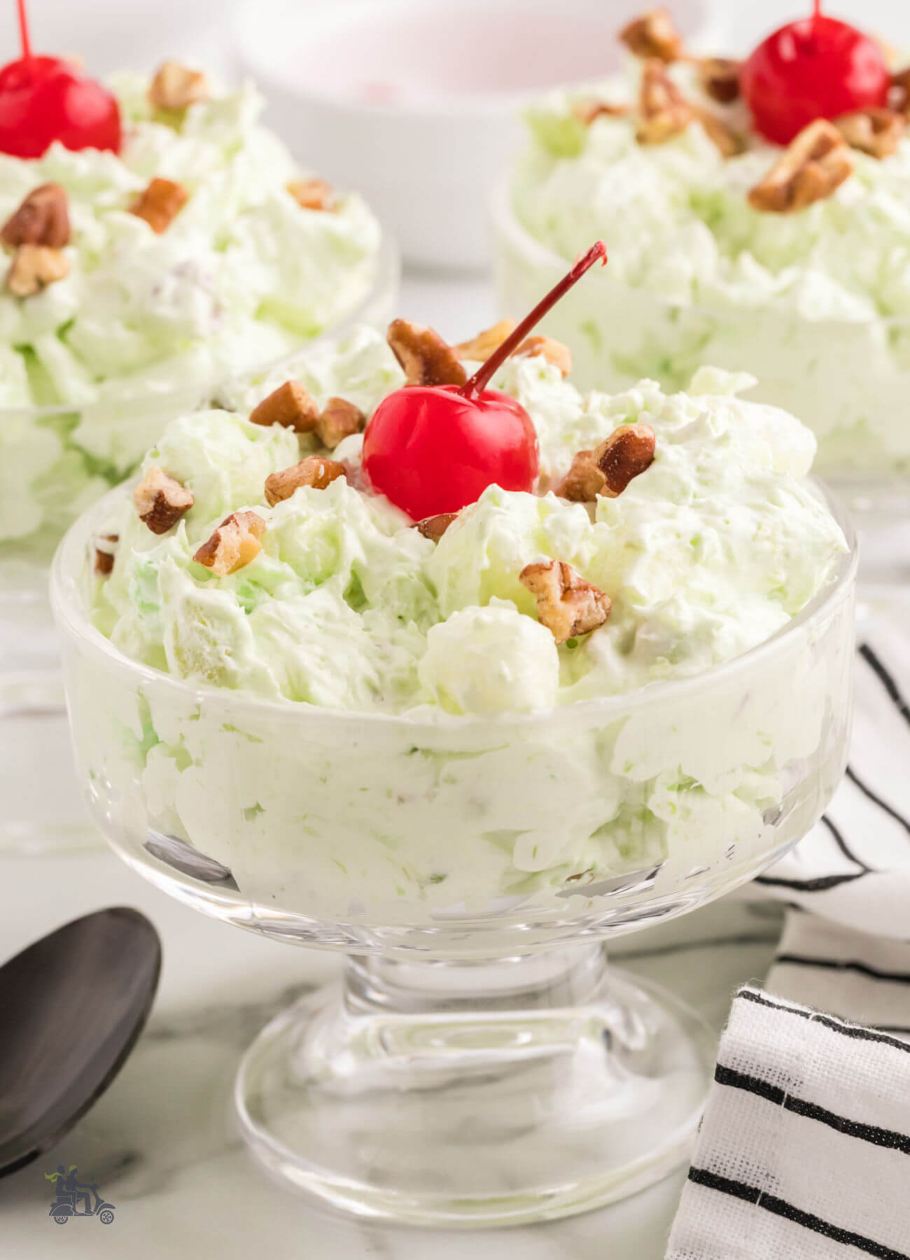 Three glass servings of Watergate Salad made with pistachio pudding and topped with a red cherry on top.