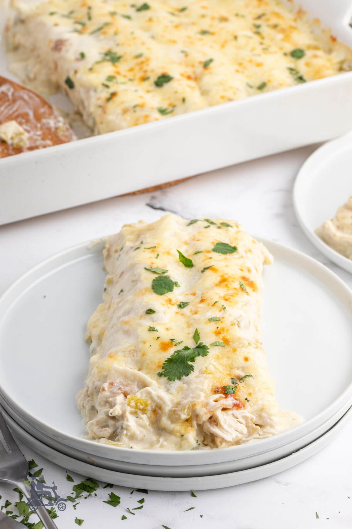 Two creamy white chicken enchiladas on a white plate with the enchilada casserole dish in the background. 