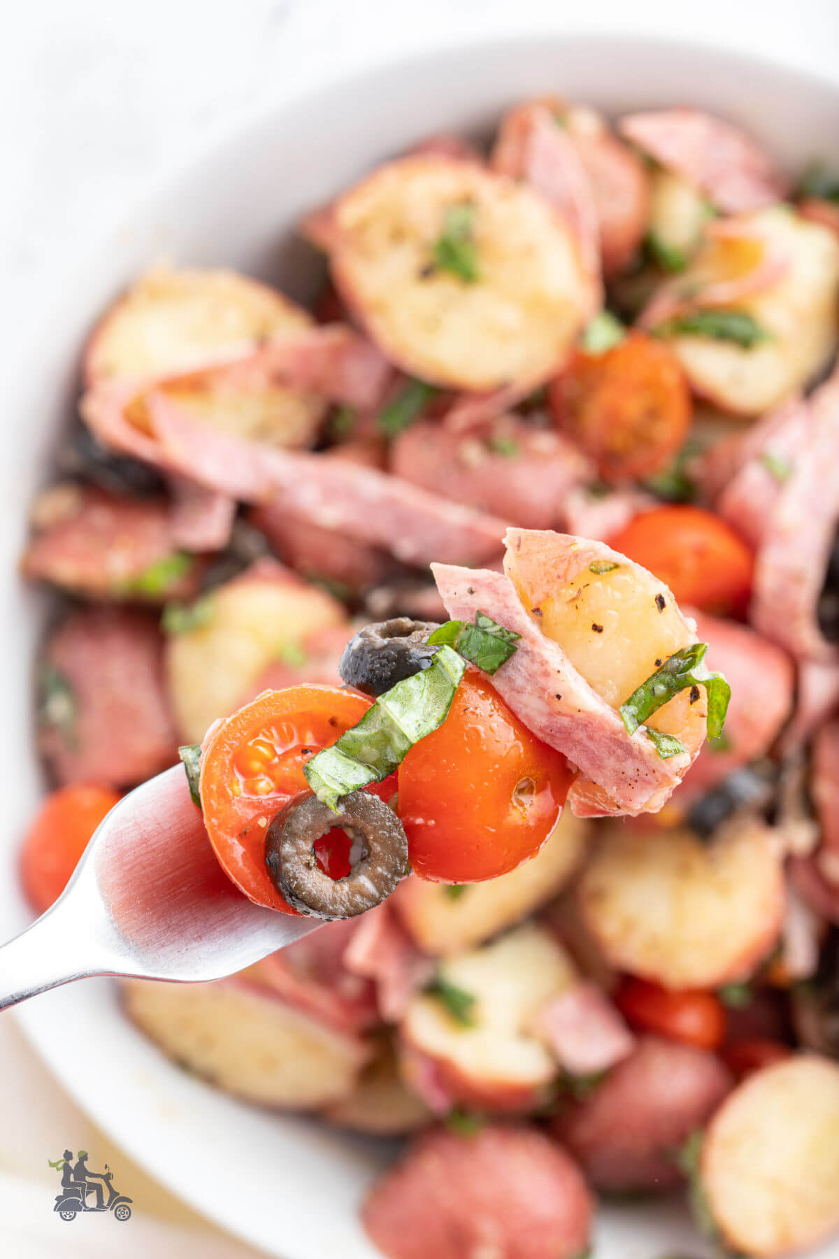 A fork holding a bite of the German like potato salad featuring Italian antipasto ingredients of black olives, salami, tomatoes, and fresh basil. 