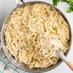 A skillet with wooden spoon mixing in the cream cheese sauce with the spaghetti and a sprinkle of chopped parsley.