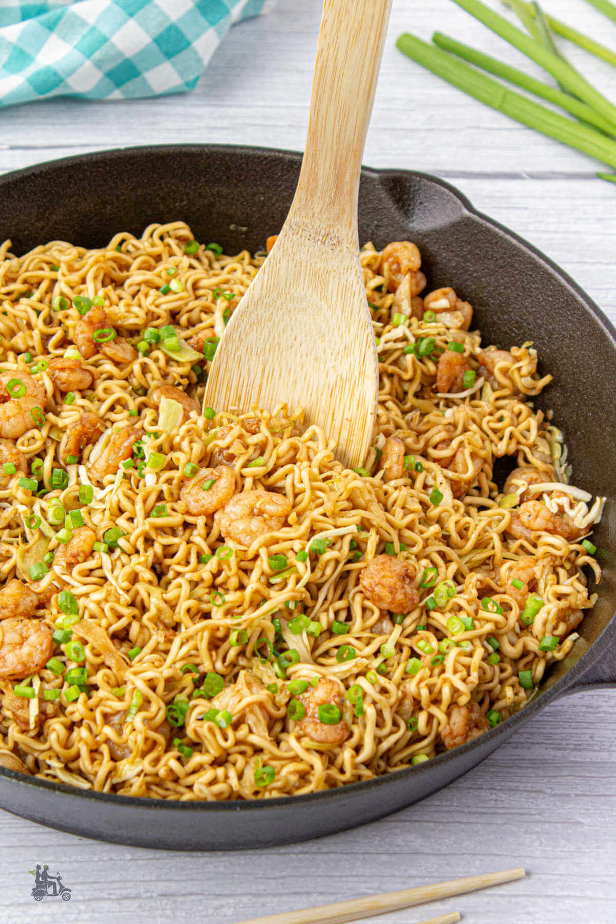 A wooden spoon in a black iron skillet ready to lift some teriyaki noodles with shrimp.