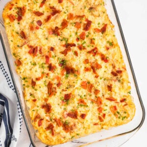 A glass casserole dish holding a recipe of loaded cauliflower bake with a serving spoon grabbing a portion.