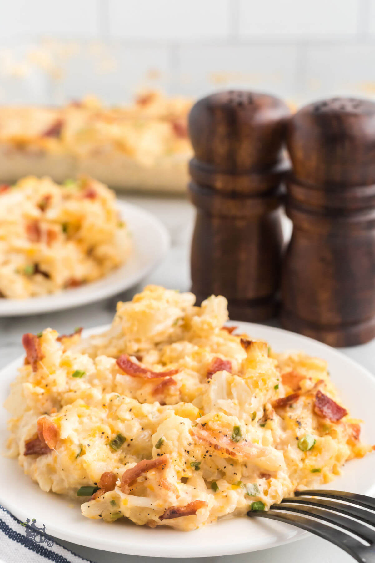 Two white plates filled with baked cheesy cauliflower casserole recipe topped with bacon. 