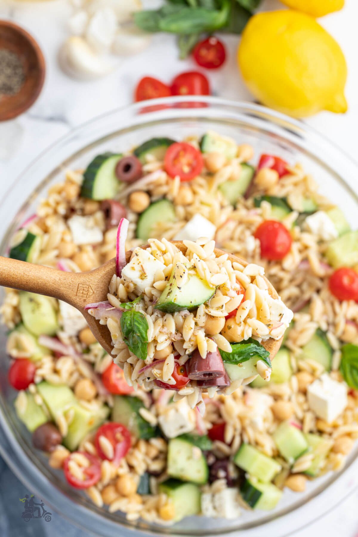Wooden serving spoon holding a spoonful of the pasta salad made with orzo pasta with Greek vegetables and seasoning. 